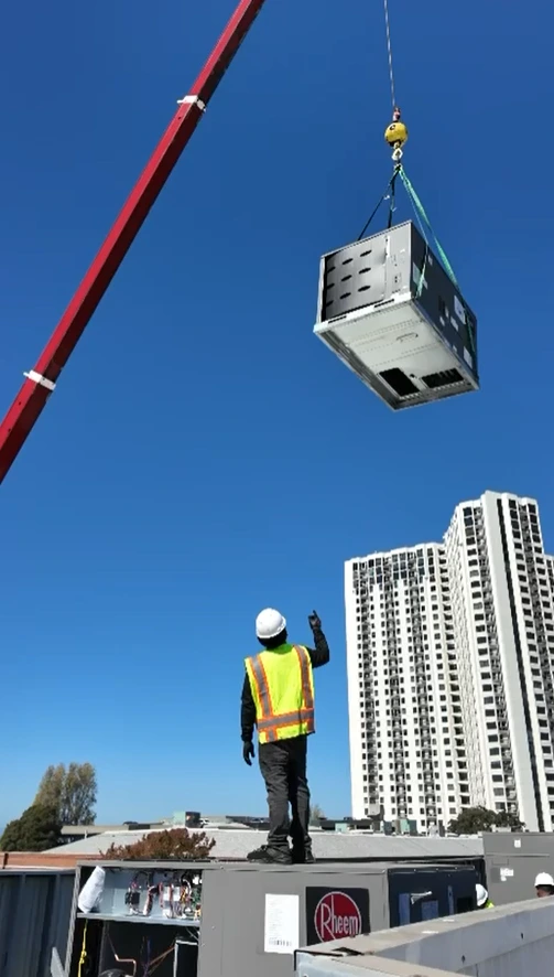 A crane lifting a packaged HVAC unit above a rooftop while a worker guides the equipment placement